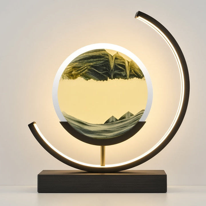 A modern table lamp with a glowing circular light inside a gold-toned frame, mounted on a black rectangular base with a curved black metal arm, set against a clean light gray background.