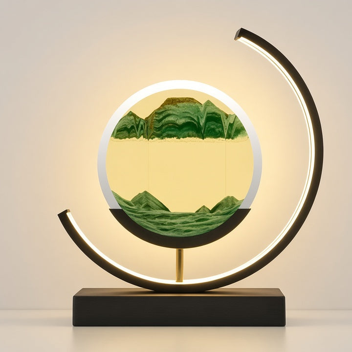 A modern LED light sculpture featuring a circular frame with a glowing warm yellow interior and a green mountain landscape design, mounted on a black rectangular base with a curved black metal arch.
